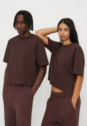 Deux personnes portant des t-shirts et pantalons amples marron foncé assortis se tiennent devant un fond clair uni, l’une avec la main derrière la tête.