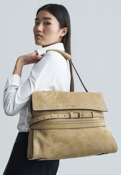 Beige suede tas met een klep, verstelbaar riemdetail en een zachte textuur. Draagtas over één schouder, gecombineerd met een wit shirt.