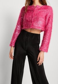 Mujer con un suéter rosa brillante de manga larga, recortado y difuso, con detalles de botones y pantalones negros de cintura alta con pliegues.