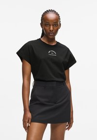 Fekete pamut póló rövid ujjakkal, fehér szöveges logóval. Egy testre szabott fekete mini szoknyával párosítva, rendezett derékpánttal és zsebekkel.