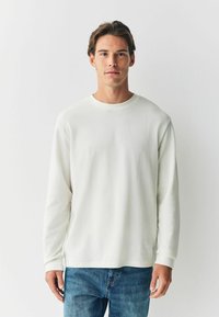 Cămașă albă cu mânecă lungă, cu un model texturat, guler rotund și croială lejeră, asociată cu blugi din denim albastru. Design minimalist.