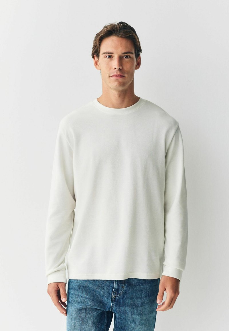Cămașă albă cu mânecă lungă, cu un model texturat, guler rotund și croială lejeră, asociată cu blugi din denim albastru. Design minimalist.