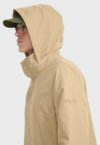 Giacca impermeabile beige con cappuccio e colletto alto. Presenta un logo discreto e una texture liscia, progettata per l'uso all'aperto.