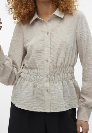 Blusa beige chiaro in tessuto testurizzato, con chiusura a bottoni sul davanti e vita elastica. Presenta maniche lunghe e un collo a camicia.