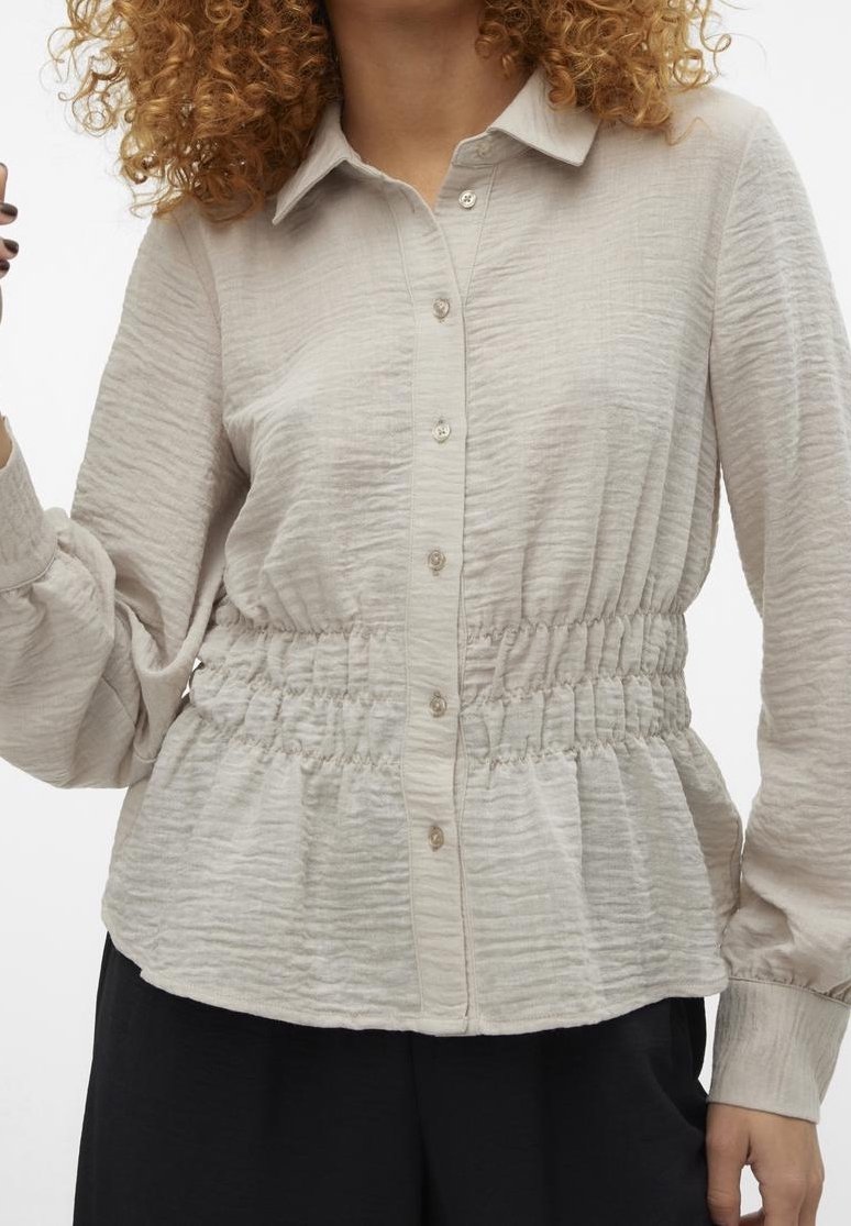 Blusa beige chiaro in tessuto testurizzato, con chiusura a bottoni sul davanti e vita elastica. Presenta maniche lunghe e un collo a camicia.