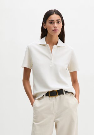 Frau mit langen dunklen Haaren, die ein weißes kurzärmliges Poloshirt, beige Hosen und einen schwarzen Gürtel trägt, steht mit den Händen in den Taschen.