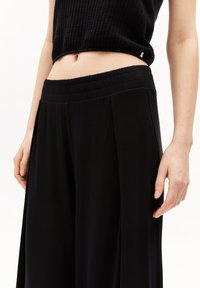 Pantalon noir à jambes larges avec une texture lisse, taille élastique et plis, présenté avec un haut noir à côtes court.