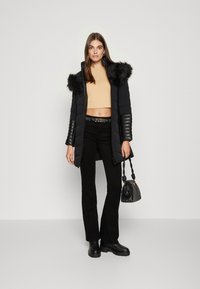 Crna puf jakna s kapuljačom od umjetnog krzna, dugi rukavi s rebrastim detaljima, uparena s bež majicom kratkih rukava, crnim širokim hlačama i malom crnom torbicom.