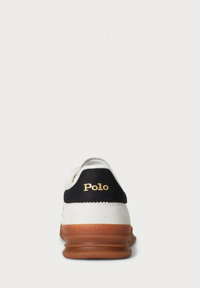 Sapatilha branca com biqueira preta e logótipo "Polo" em dourado. Apresenta um tecido texturizado na parte superior e sola em borracha em tom castanho claro.