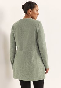 Lang grøn cardigan lavet af ribstrikket stof, med en tætsiddende talje og lange ærmer, med en rund halsudskæring og en let udvidet kant.