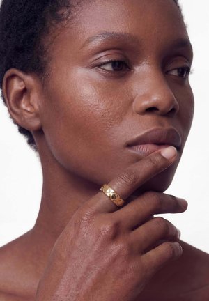 Primer plano de una persona de piel oscura tocándose el mentón con una mano que lleva un anillo dorado con estampado, mirando pensativamente hacia un lado.