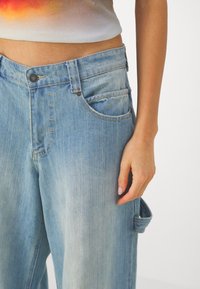 Světle modré džíny s volným střihem, které mají přední kapsy, viditelné švy, zapínání na knoflík a postranní smyčku.