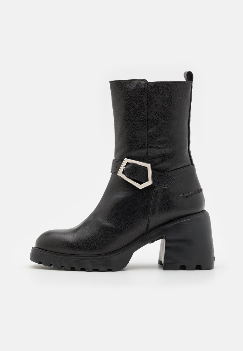 Botte en cuir noire mi-mollet avec talon bloc épais, semelle crantée et bride décorative avec boucle argentée sur le côté extérieur.