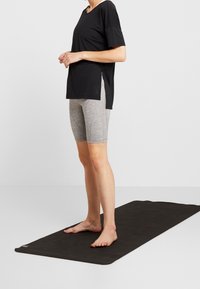 Camiseta negra de corte holgado con mangas cortas, combinada con pantalones cortos de ciclismo gris claro. De pie sobre una alfombra negra, descalzo, frente a un fondo liso.