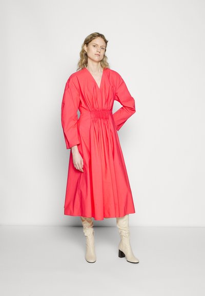 Vestido longo de mangas compridas na cor rosa coral, com decote em V e cintura franzida, apresentando uma saia evasê. Um par de botas de cano alto em creme, com salto, completa o visual.