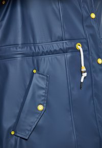 Tamsiai mėlynas vandeniui atsparus švarkas su lygiu paviršiumi, priekinė kišenė, geltoni mygtukai-užsegimai ir reguliuojama raištelė liemens srityje.