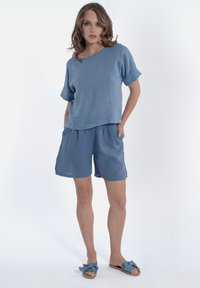 Top in lino blu con maniche corte, abbinato a pantaloni corti larghi dello stesso colore. Gli accessori includono sandali blu con fiocco. Tessuto liscio, design casual.