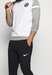 Pullover con corpo bianco e maniche grigie, caratterizzato da un logo rotondo, abbinato a pantaloni tuta neri con logo Nike bianco, vestibilità slim.