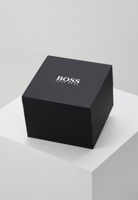 Juodas dovanų dėžutė su lygiu paviršiumi. Viršuje yra balta logo, kuri skaitoma "BOSS HUGO BOSS", centruota ant viršaus.