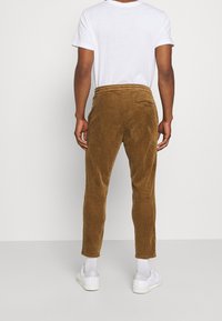 Persoon in bruine corduroy broek, wit T-shirt met korte mouwen, witte sokken en witte Adidas sneakers, staand met de rug naar de camera tegen een effen achtergrond.