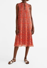 Robe sans manches à motif rouge avec un décolleté en goutte d'eau, texture plissée et silhouette fluide, associée à des sandales à lanières marron.
