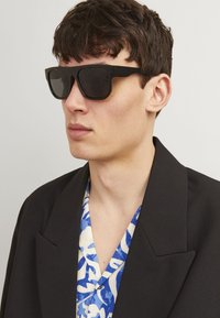 Schwarze quadratische Sonnenbrillen mit großen Gläsern und dicken Rahmen. Getragen von einer Person in einem schwarzen Blazer über einem blau-weiß gemusterten Hemd.
