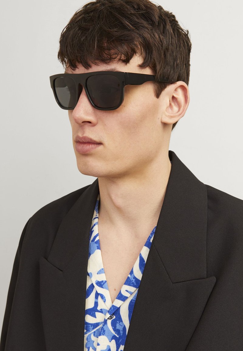 Schwarze quadratische Sonnenbrillen mit großen Gläsern und dicken Rahmen. Getragen von einer Person in einem schwarzen Blazer über einem blau-weiß gemusterten Hemd.