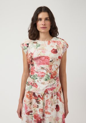 Femme aux cheveux foncés mi-longs portant une robe sans manches à motif floral dans des tons rose, rouge et vert sur fond blanc.