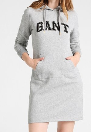 Femme portant une robe sweat à capuche gris clair avec le logo "GANT" sur la poitrine, mains dans la poche avant.