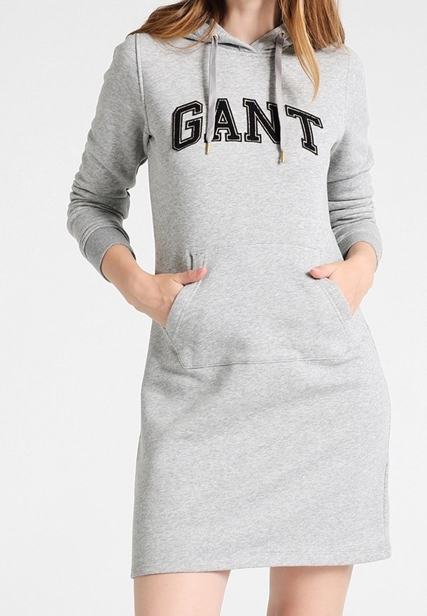 Femme portant une robe sweat à capuche gris clair avec le logo "GANT" sur la poitrine, mains dans la poche avant.