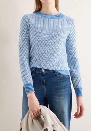 Person wearing a light blue textured sweater and blue jeans holding a beige jacket in one hand against a plain background.