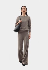 Jasnobrązowy dzianinowy sweter oraz pasujące szerokie spodnie z prążkowanym dekoltem i mankietami, zestawione z czarną torebką i butami.