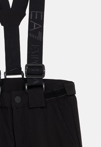 Čierne nohavice s nastaviteľnými trakovými popruhmi s prackou a jemným textom, a pásom s gombíkom a suchým zipsom.