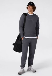 Grauer Sweatshirt mit gesticktem Logo, passende graue Jogginghose, schwarzer Rucksack und graue Sneakers. Kappe mit Logo und sichtbares lockiges Haar.