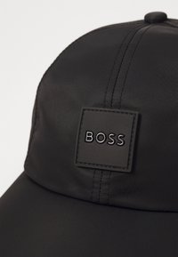 Gorra negra hecha de tela suave, con un parche rectangular texturizado que lleva el logotipo "BOSS" en letras negras. Visera curva y detalles de costura visibles.