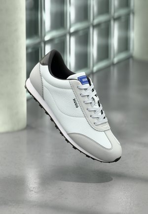 Valge ja halli nahast toss musta kannaga ja valge paeltega, kaubamärgiga „HUGO“, hõljuv siseruumides üle halli läikiva põranda.