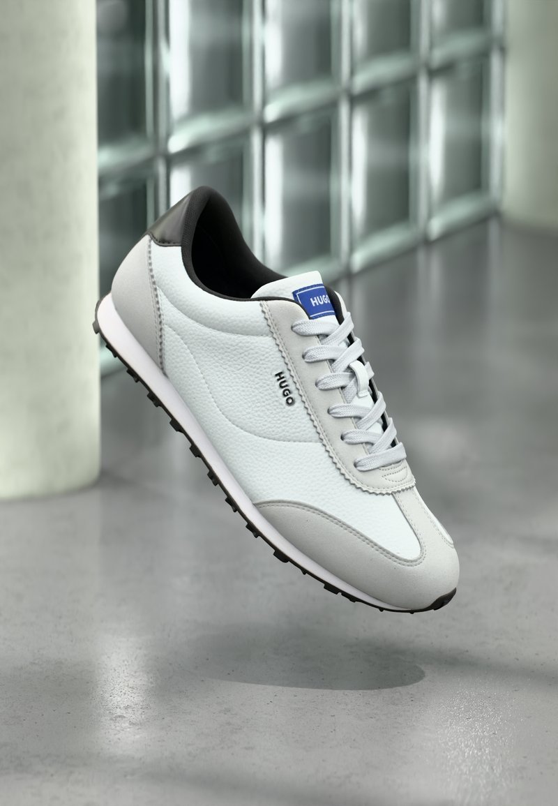 Baskets en cuir blanc et gris avec talon noir et lacets blancs, marque "HUGO", flottant au-dessus d'un sol gris poli à l'intérieur.