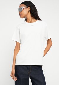 Balta kokvilnas T-krekls ar īsām piedurknēm, krūšu daļā neliels sudraba logotips. Kombinēts ar tumši ziliem džinsiem un caurspīdīgiem saulesbrillēm.