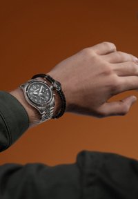 Sølvur med sort urskive og nummeriske markeringer på et metalarmbånd, parret med et sort flettet armbånd, der viser et lille rundt ur.