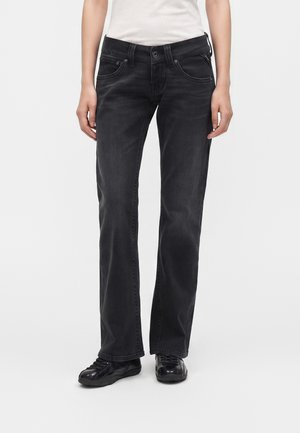 Personne portant un jean noir droit, une chemise blanche et des baskets noires, debout devant un fond blanc uni.