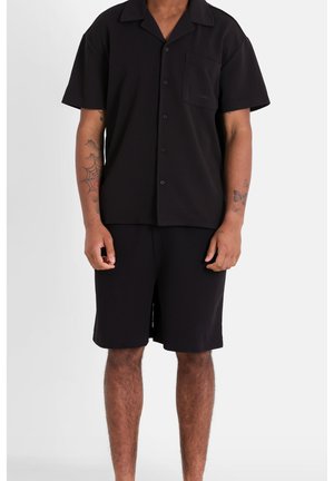 Camisa negra de manga corta con bolsillo y cierre de botón, combinada con pantalones cortos negros. Tela suave, corte relajado, sin patrones ni acentos visibles.