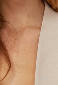 Beżowy, teksturowany materiał układa się na obojczyku, odsłaniając odcień skóry. Cienkie pasemka włosów widoczne są na gładkiej powierzchni.
