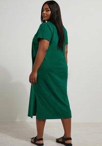 Zielona, sięgająca do kolan sukienka z miękkiej bawełny, z krótkimi rękawami i bocznymi rozcięciami dla łatwości ruchu. Noszona z czarnymi sandałami.