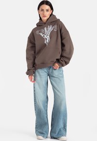 Brązowa bluza z kapturem z graficznym wzorem dłoni w białym kolorze, w połączeniu z jasnoniebieskimi szerokimi spodniami dżinsowymi i kremowymi sneakersami.