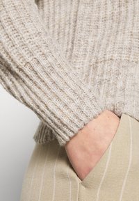 Personne portant un pull en maille côtelée beige avec la main dans la poche d'un pantalon beige à fines rayures, mettant en valeur les textures et les détails du tissu.