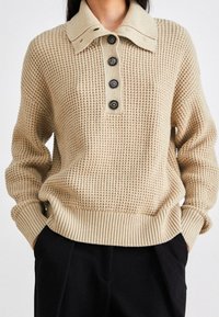 Beige wafelbrei trui met vier zwarte knopen, opstaande kraag, afgeknipte mouwen en geribde zoom, gedragen met zwarte broek.