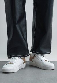 Hvite lave sneakers med beige semsket skinn detaljer, brukt med svarte vide bukser på grått gulv bakgrunn.