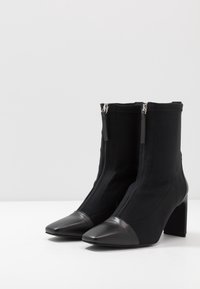 Bottines noires avec un dessus en textile et un bout en cuir pointu. Présente des doubles zips et un large talon bloc. Texture lisse et design épuré.