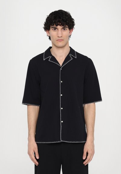 Camisa negra de manga corta con costuras blancas contrastantes en el cuello y los puños, con un botón en la parte delantera. Textura de tela suave.