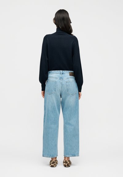 Μπλε πουλόβερ με υψηλό γιακά και χαμηλωμένους ώμους, συνδυασμένο με ανοιχτόχρωμα τζιν φαρδιάς γραμμής και παπούτσια με λεοπάρ μοτίβο.